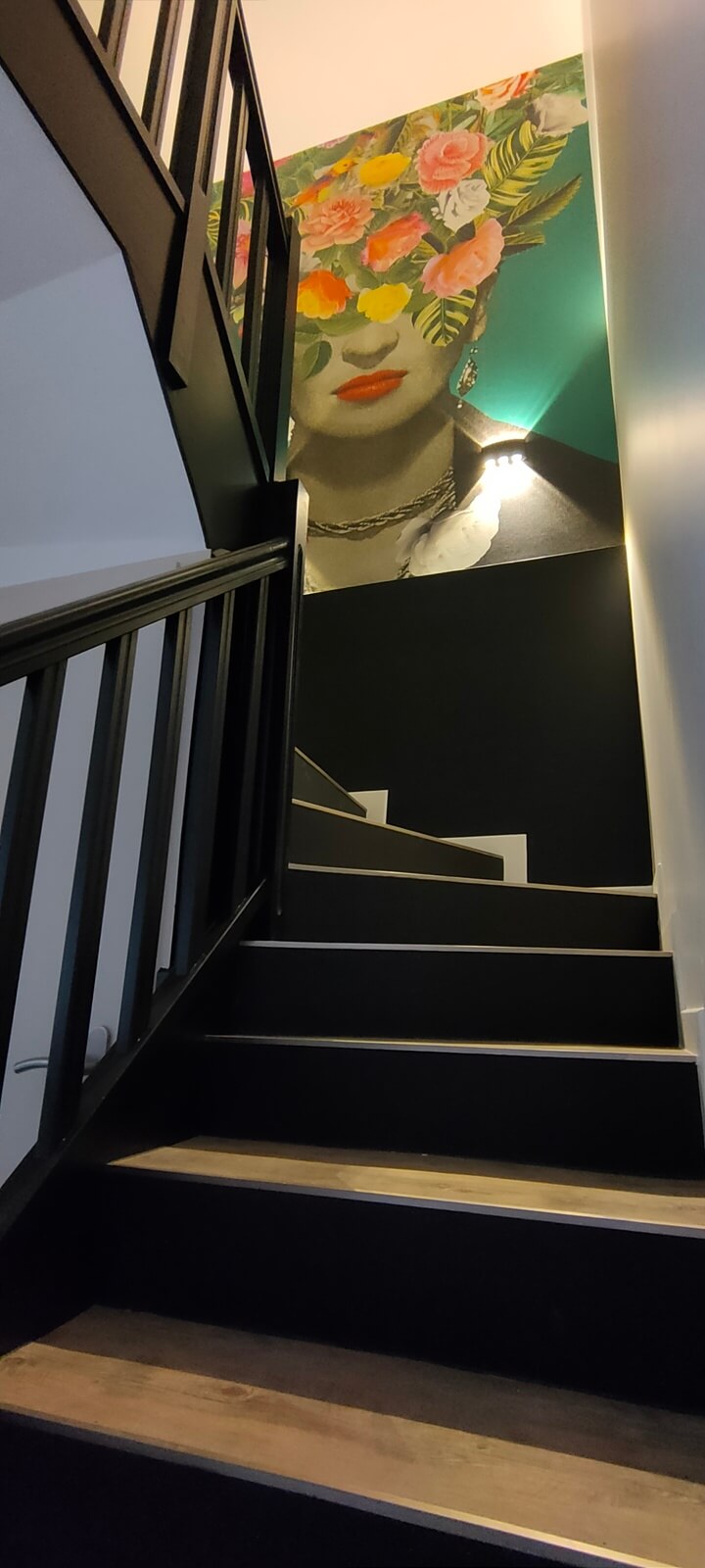 Une montée d'escalier avec des marches en bois, une rampe peinte en noir, des murs blancs et noirs décorés d'un tableau Frida Kahlo après home staging