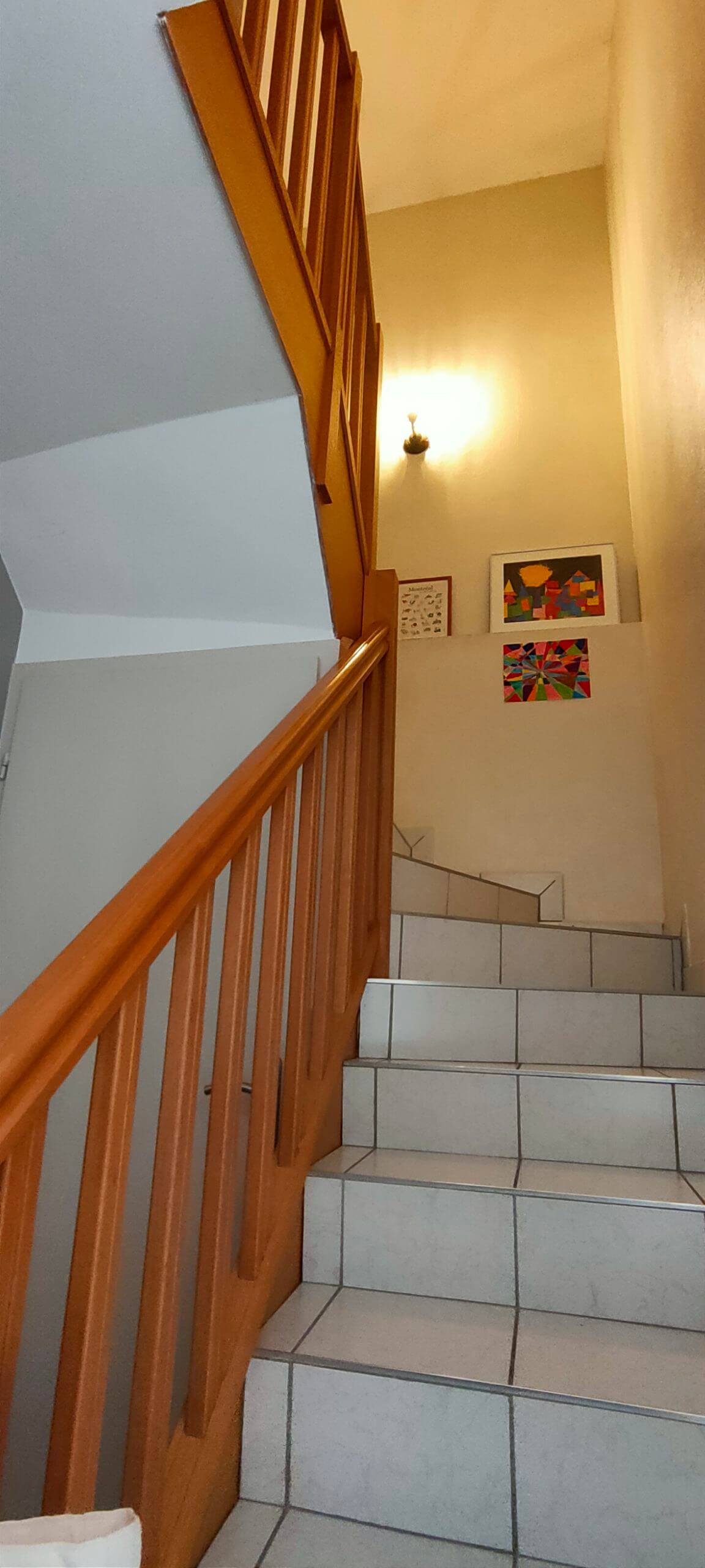 Une montée d'escalier avec des marches en carrelage blanc, une rampe couleur chêne et des murs jaunes clairs avant un home staging